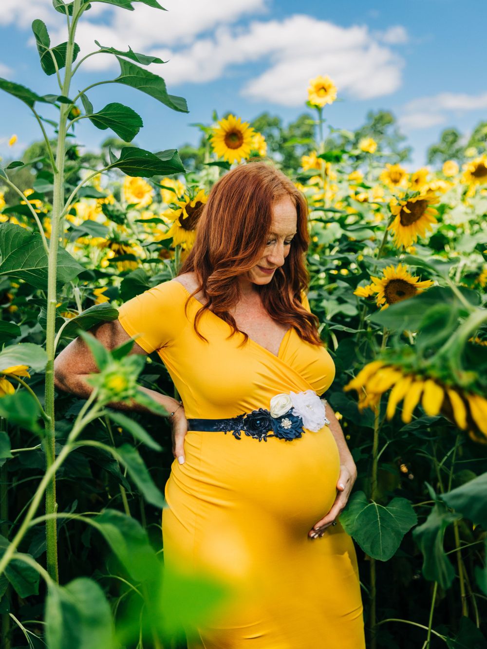 A maternity photo of someone in a yellow dress standing among blooming sunflowers.