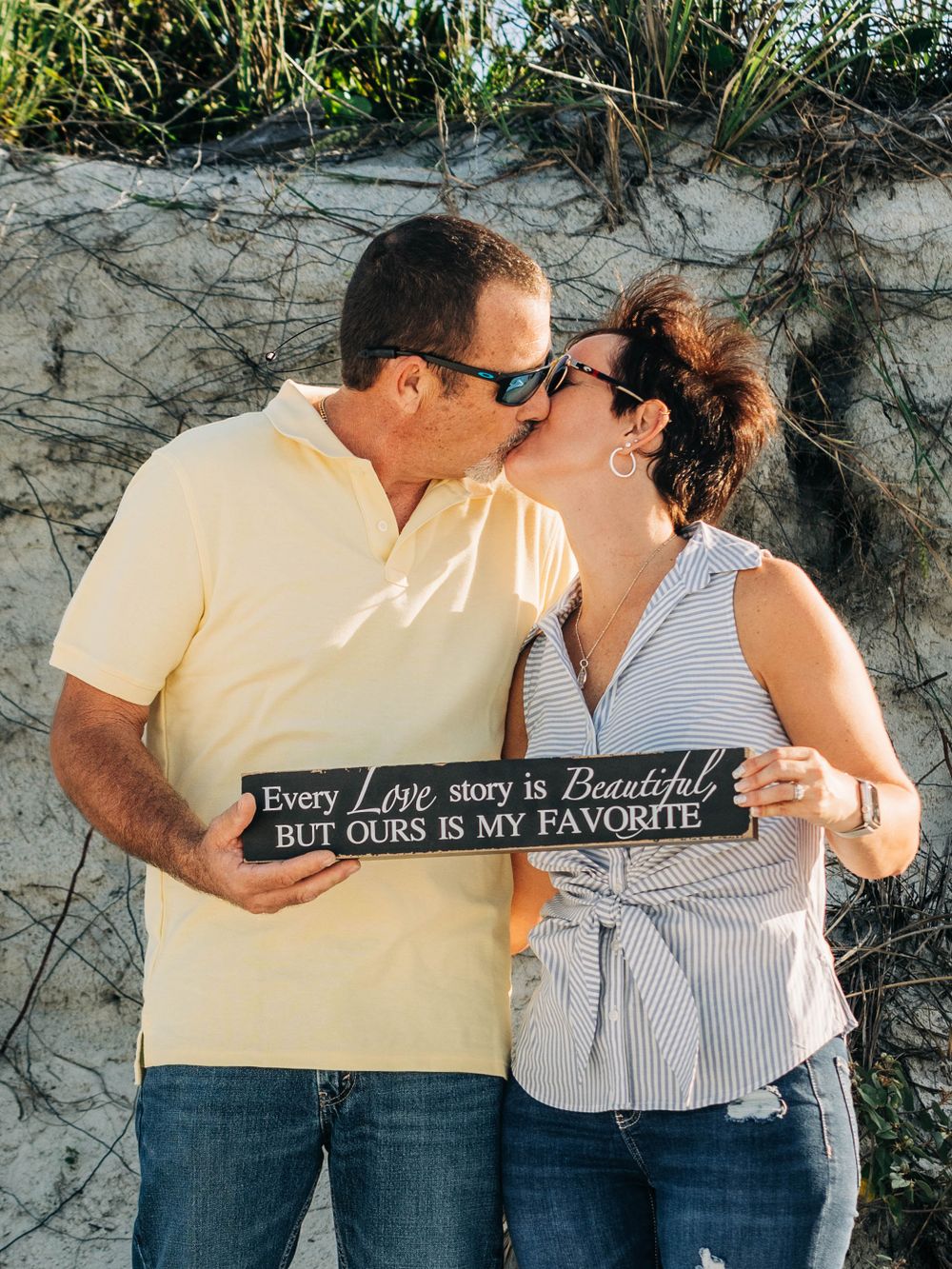 A couple sharing a romantic moment on the beach while holding a sign about love stories.