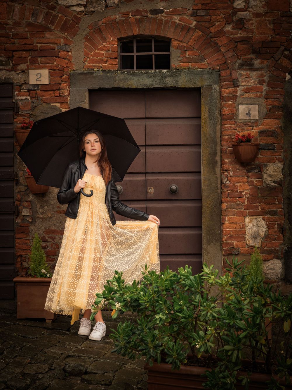 Figure in leather jacket and yellow dress poses with umbrella by rustic doorway.