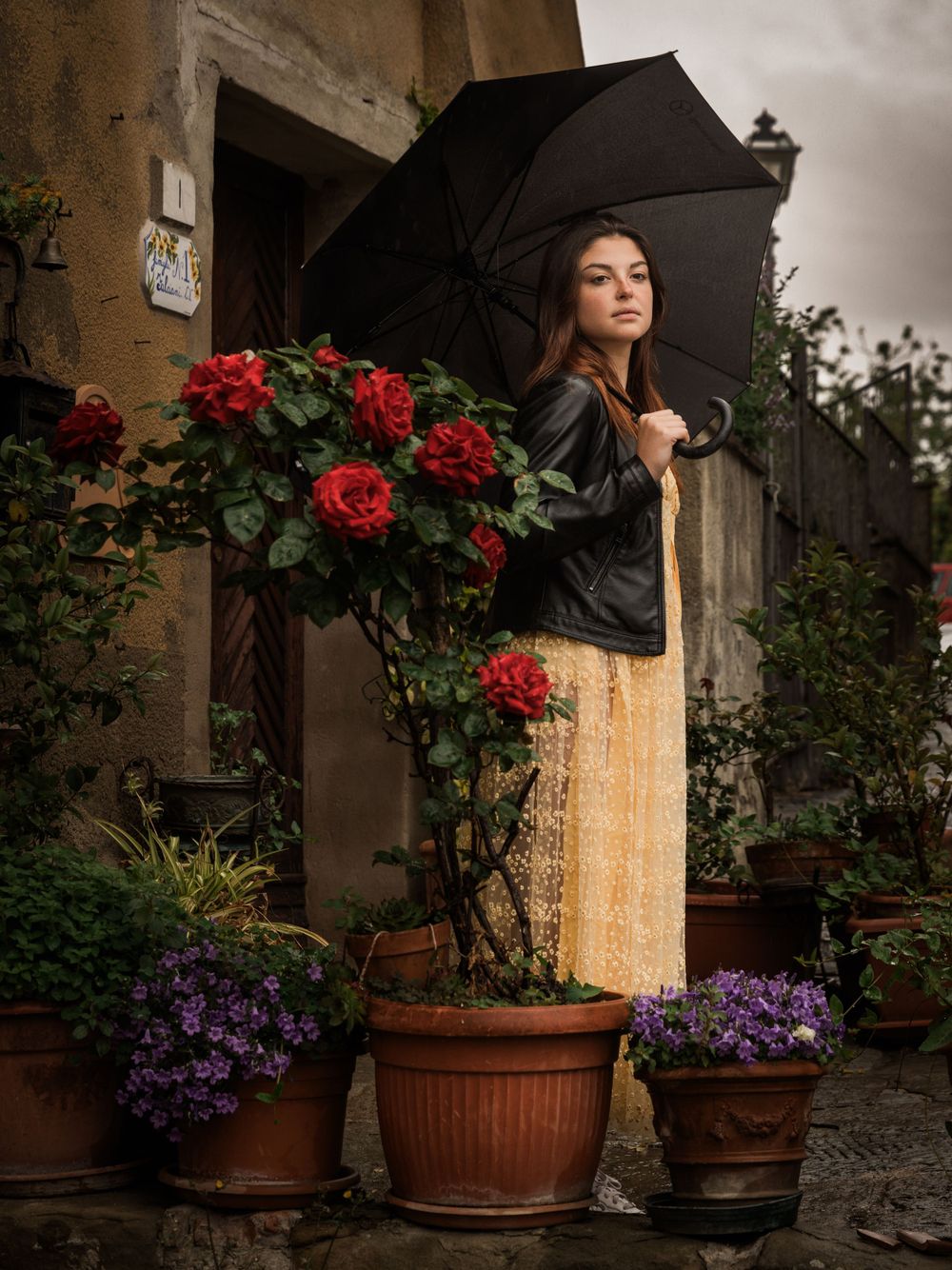 Person with umbrella standing beside rose bush and potted flowers against stone wall.