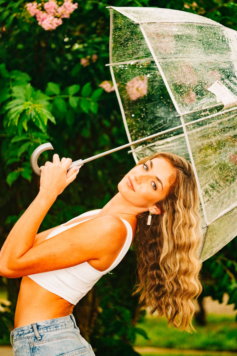 Summer portrait with clear umbrella and casual outfit against lush garden setting.