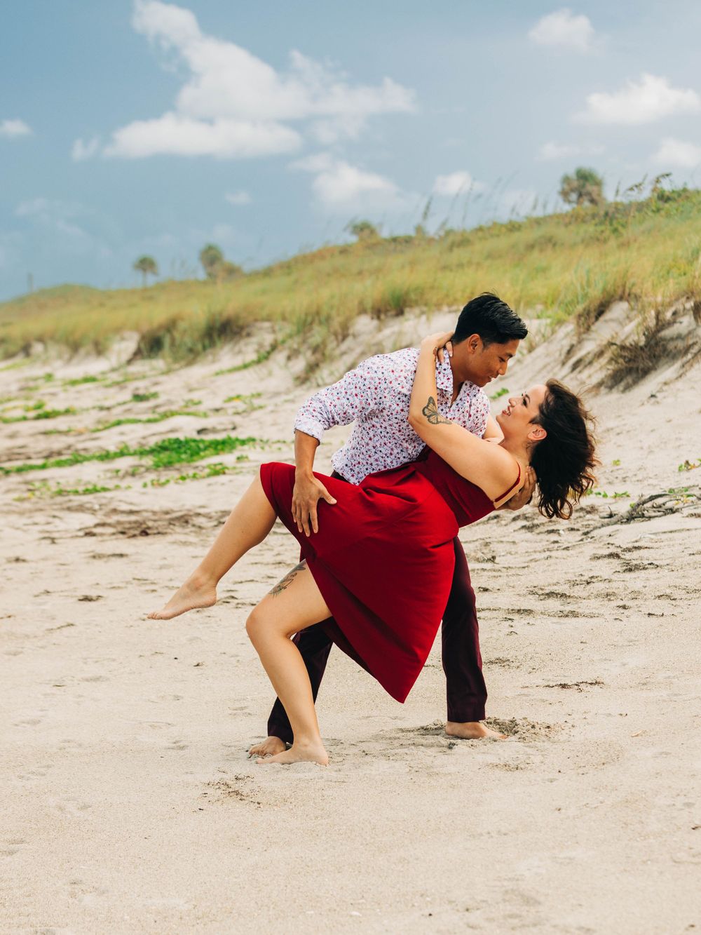 Romantic couple dancing on beach, with one wearing red dress and other in patterned shirt.