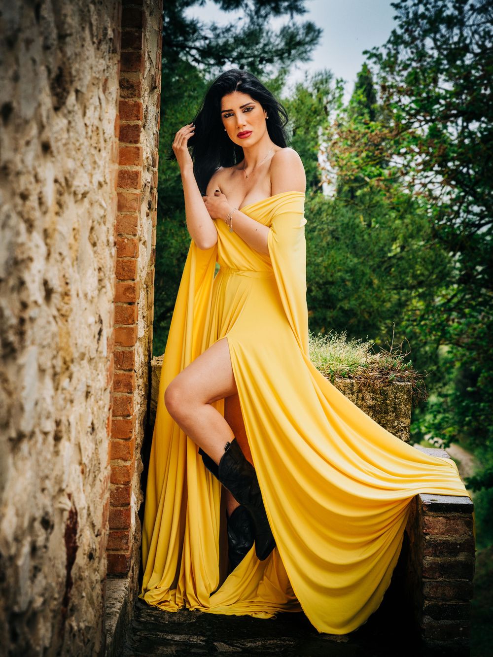 Dramatic fashion photo featuring flowing yellow dress against stone wall and greenery.