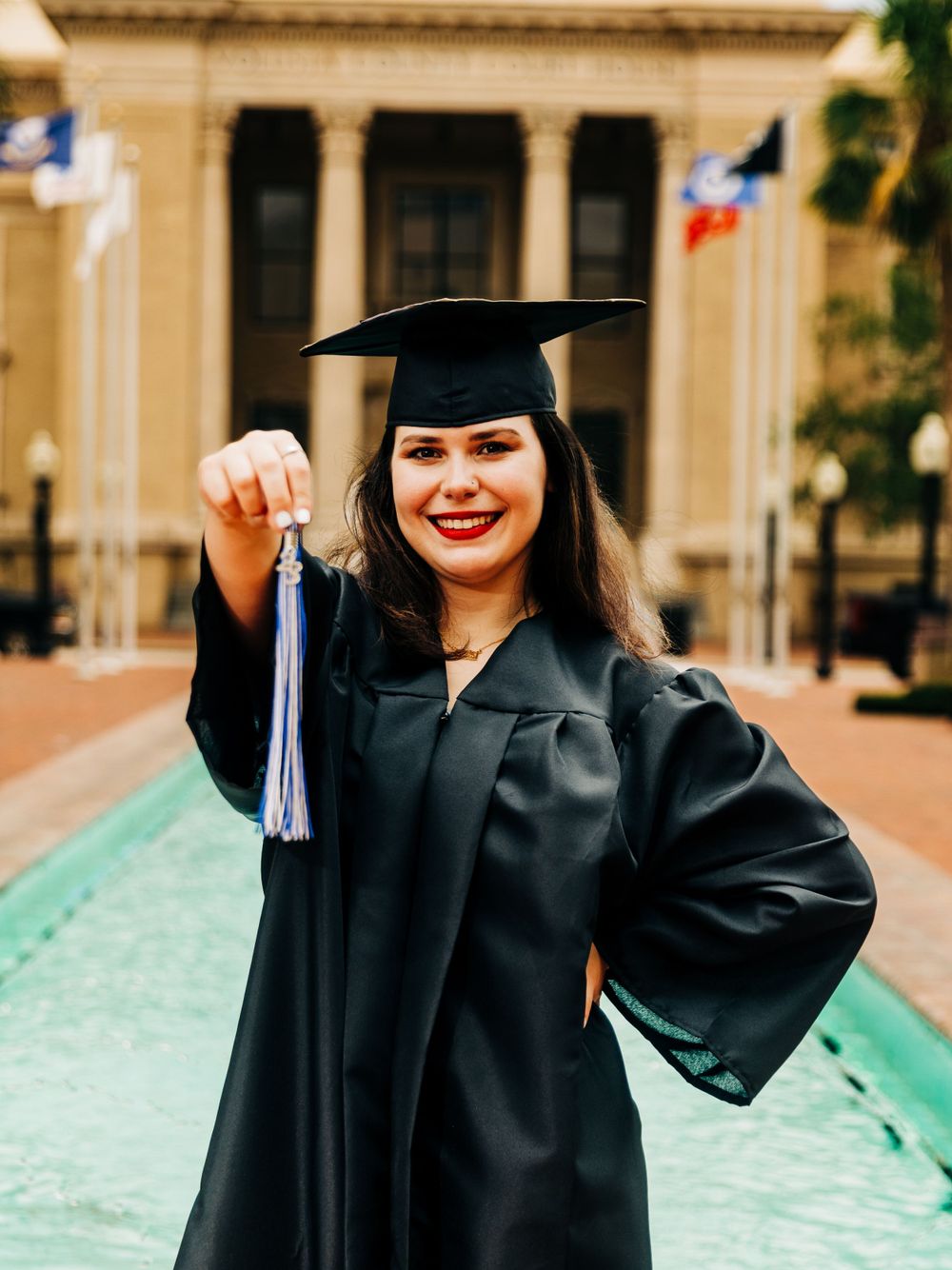 A graduate in cap and gown holds their tassel on a campus walkway.
