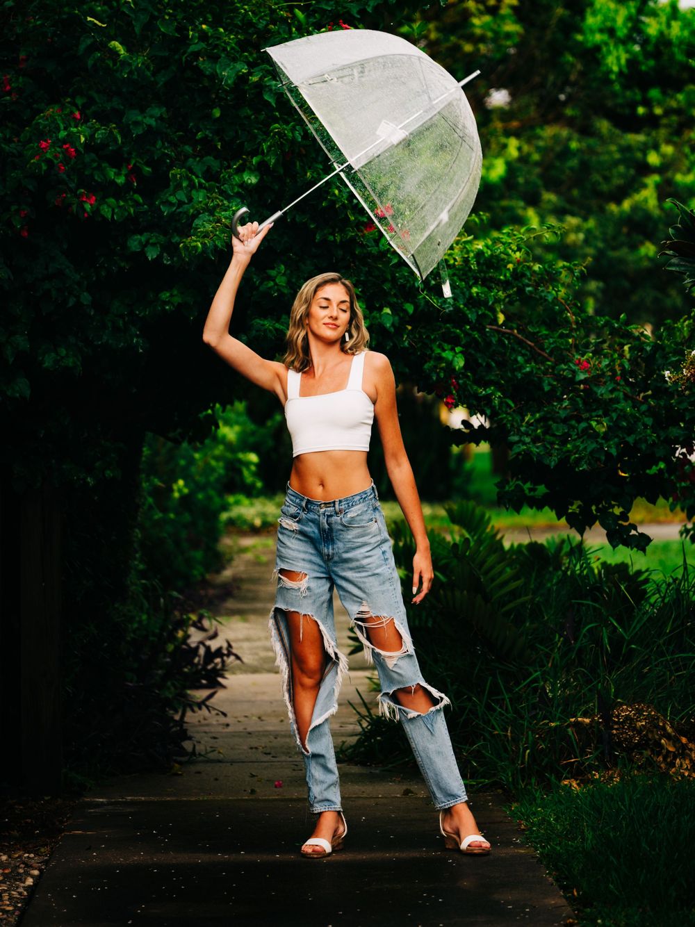 Candid outdoor shot with clear umbrella, white crop top and distressed denim on garden path.