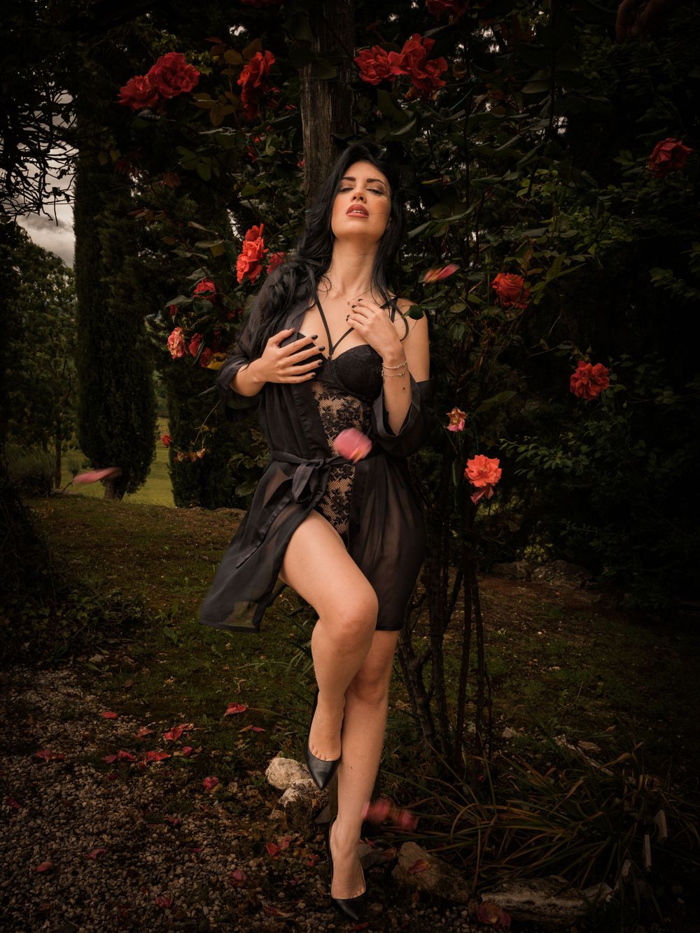 Artistic fashion photo in black dress surrounded by red roses in garden setting.