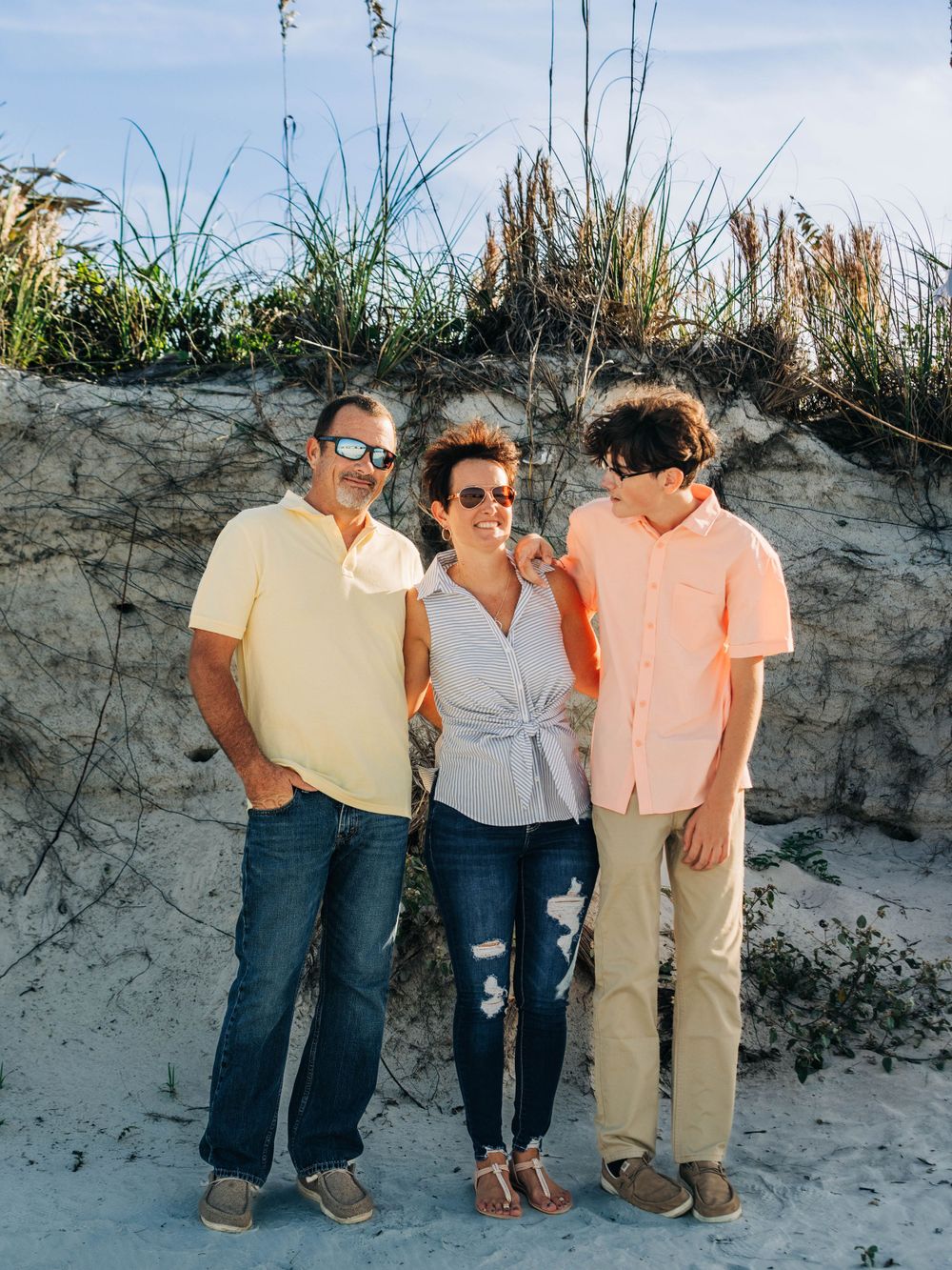A family of three posed together on a sandy beach dune during golden hour.