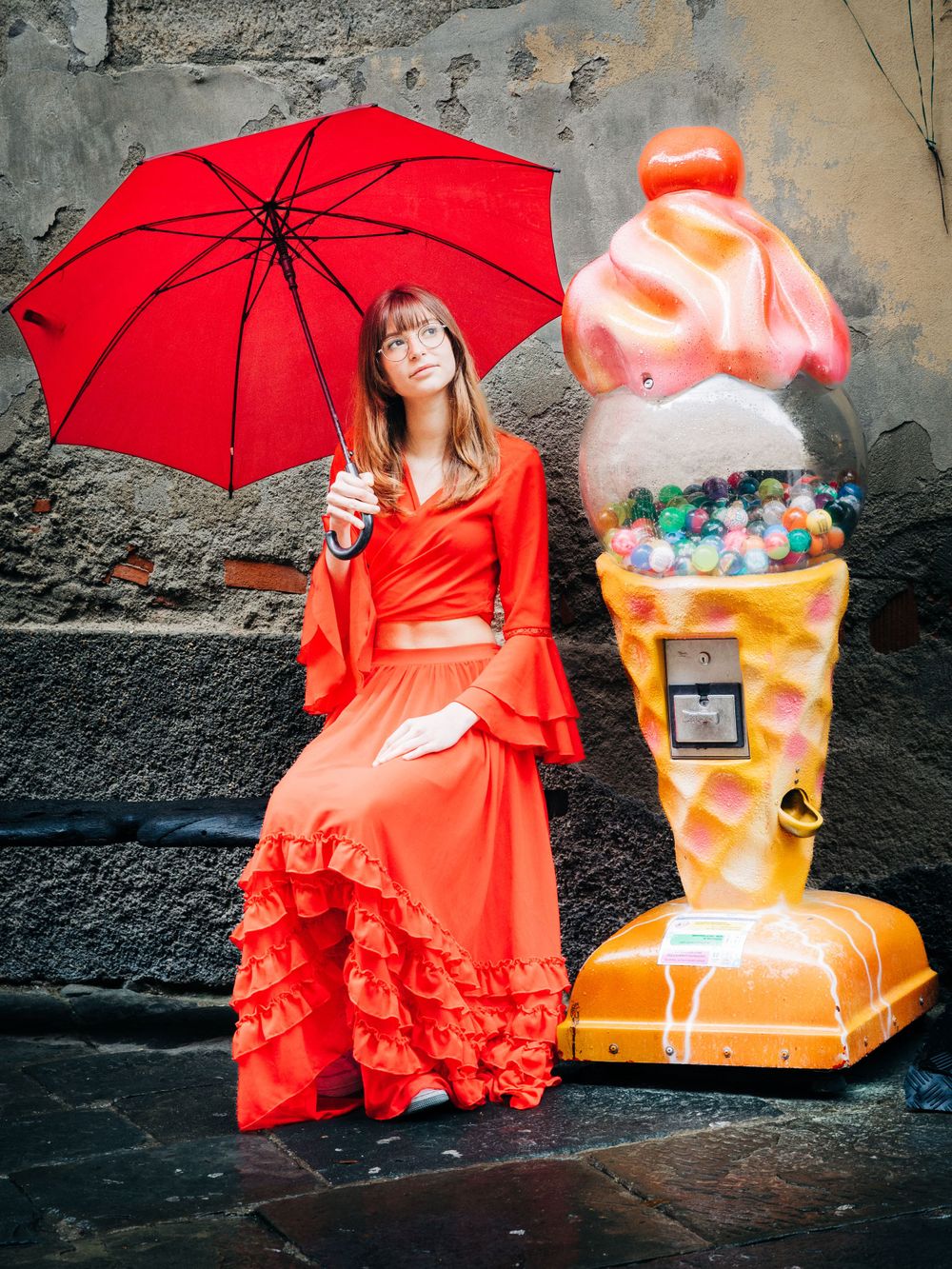 Woman in flowing red dress poses with umbrella next to large ice cream sculpture decoration.