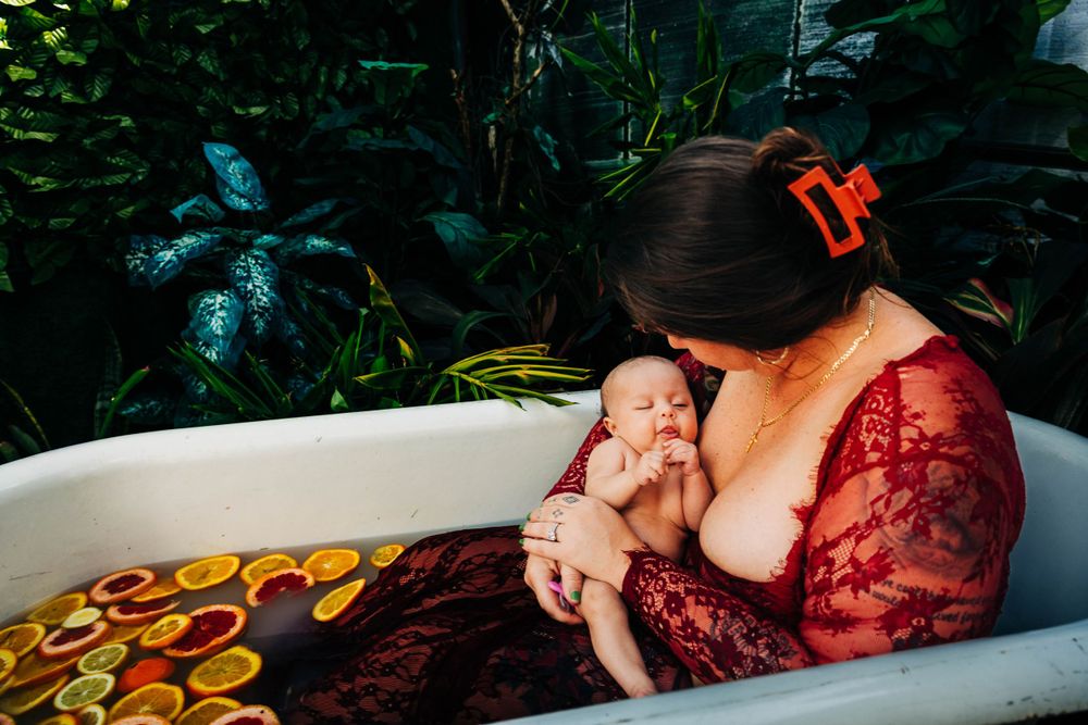A tender nursing moment captured in a bathtub surrounded by lush greenery and floral patterns.