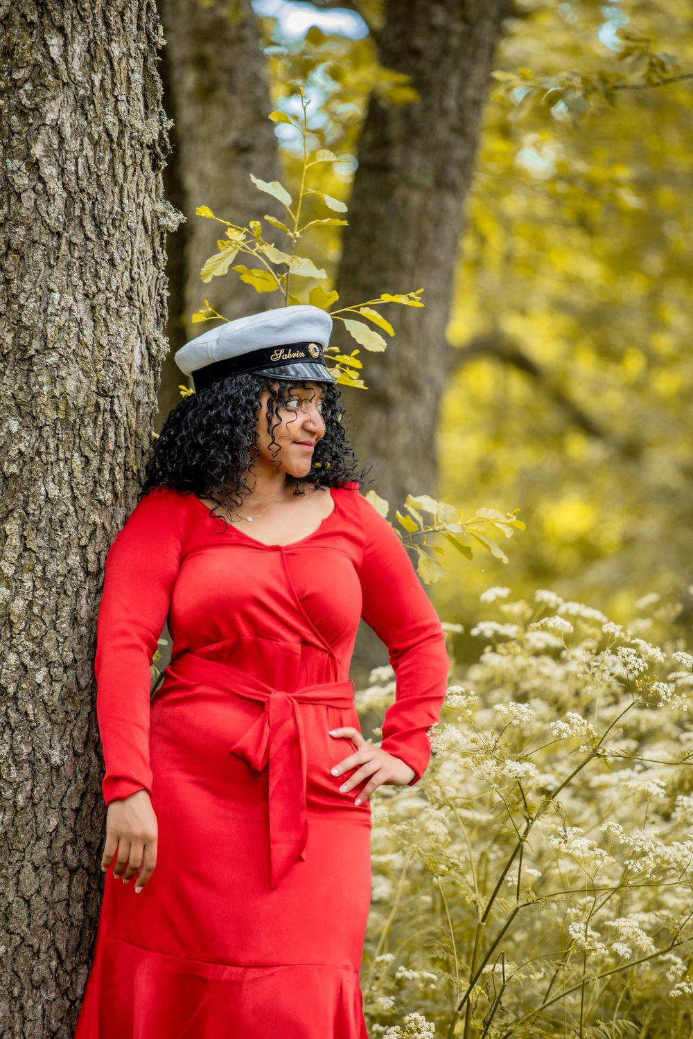 Person in red dress and hat poses elegantly against tree trunk in autumn setting.