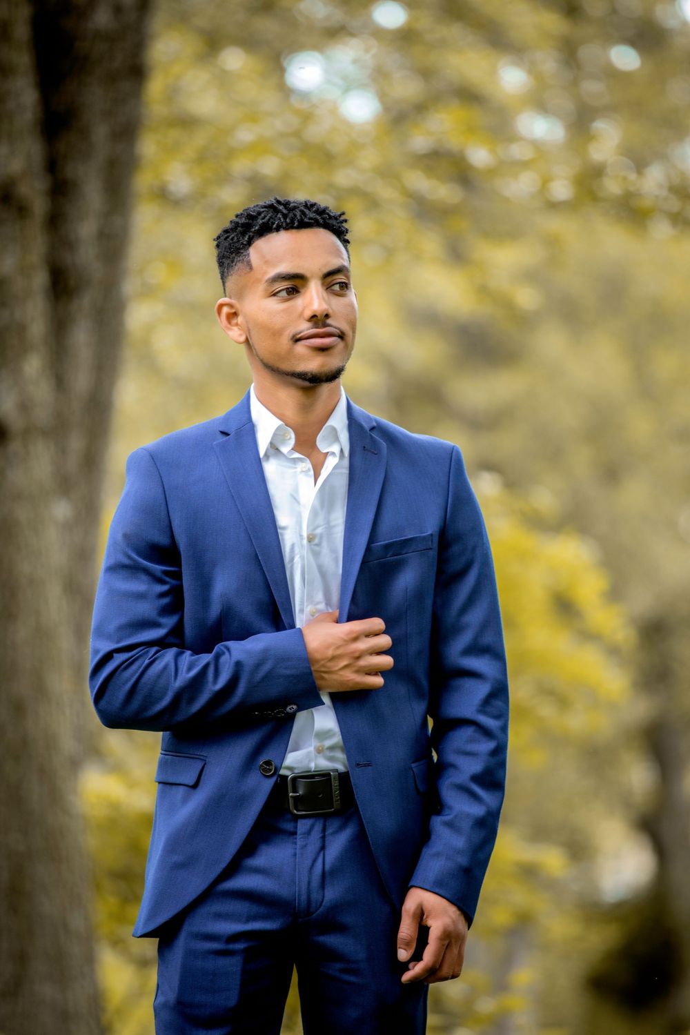 Professional wearing navy blue suit stands confidently in outdoor autumn setting.