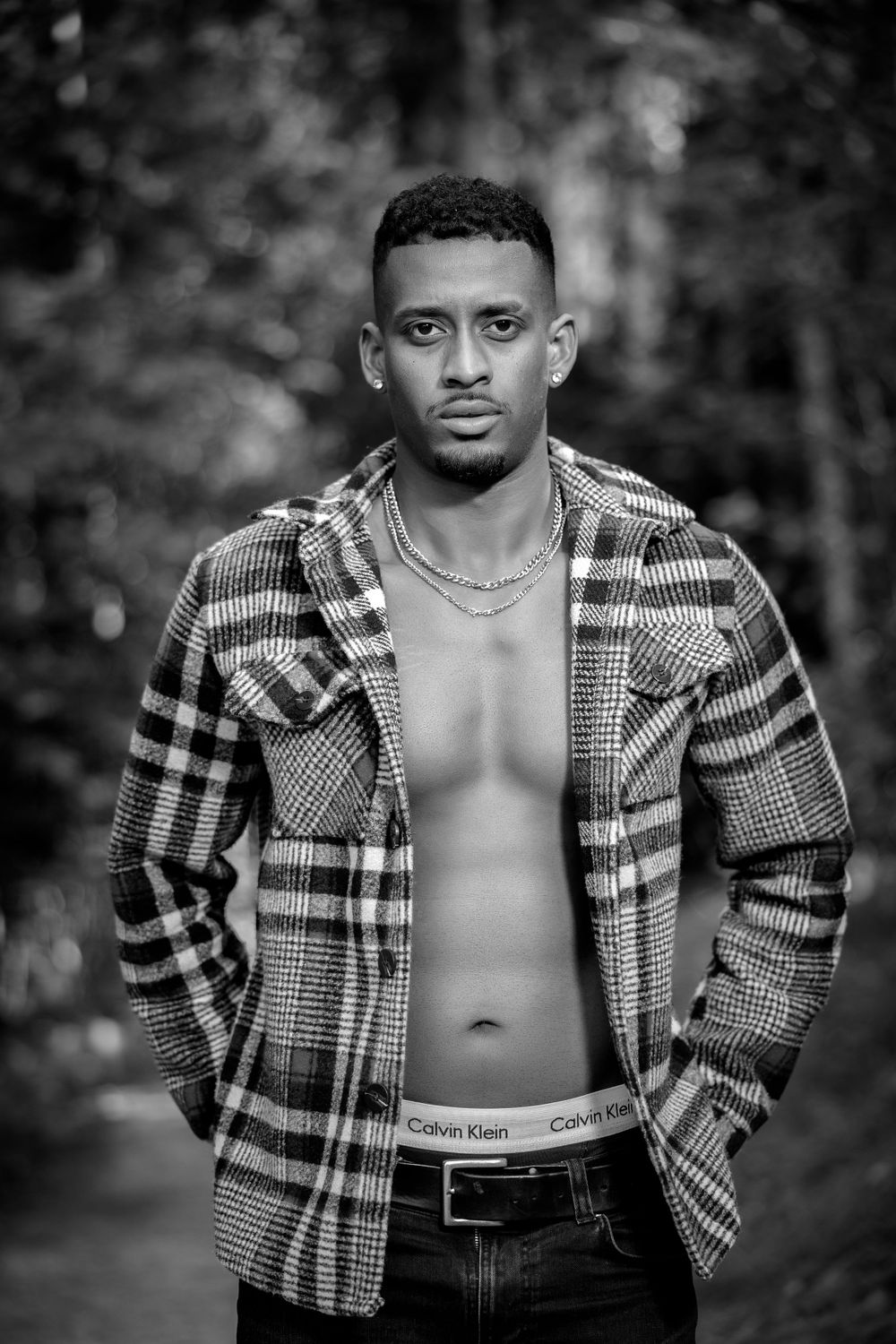 Dramatic black and white portrait of model in plaid jacket against forest backdrop.