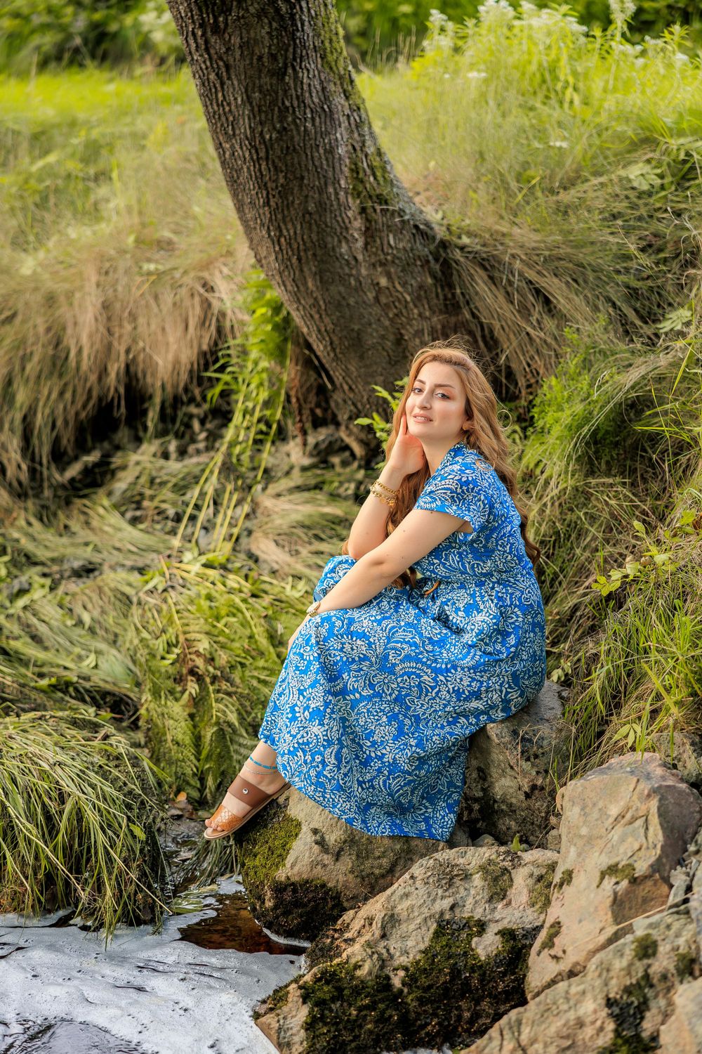 Person in flowing blue dress sits gracefully on rocks by water's edge in natural setting.