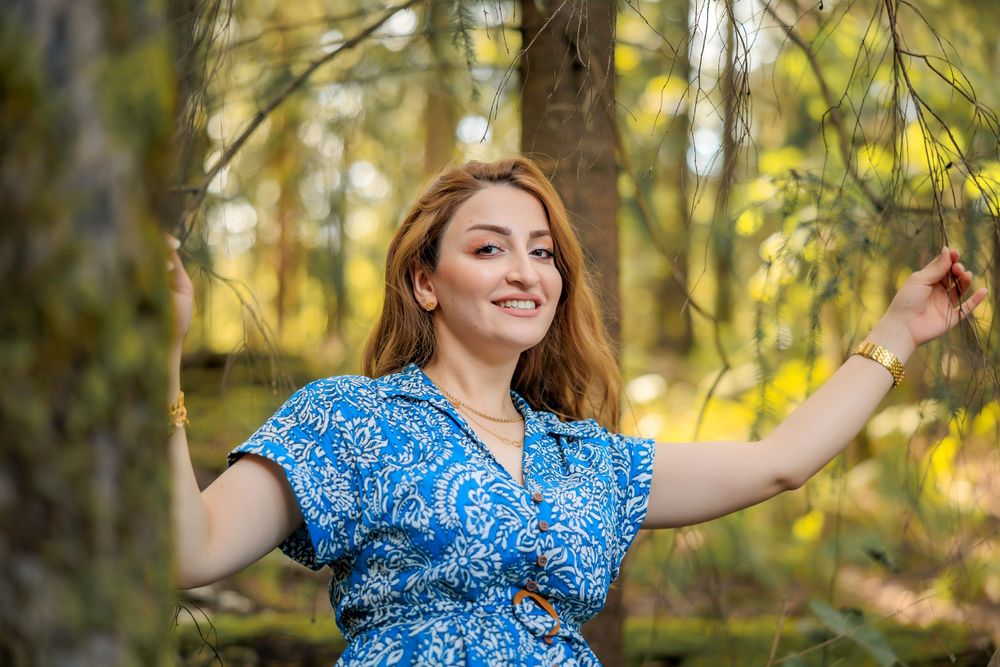 Person wearing blue floral dress dances playfully in a sunlit wooded area.