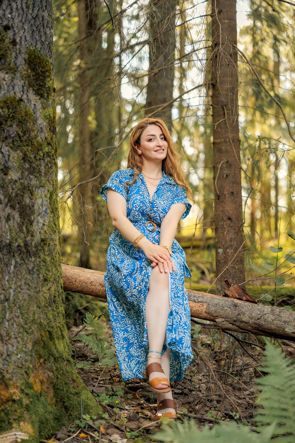 Woman in patterned blue dress sits gracefully on fallen log in forest.