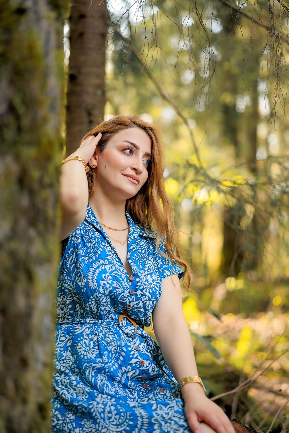 Woman in blue patterned dress poses thoughtfully against blurred forest background in natural lighting.