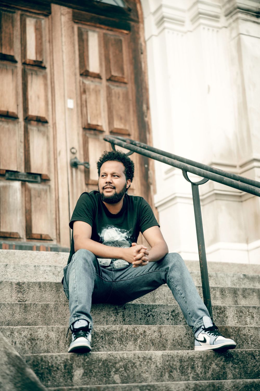 Candid portrait on church steps wearing black t-shirt and jeans with architectural backdrop.