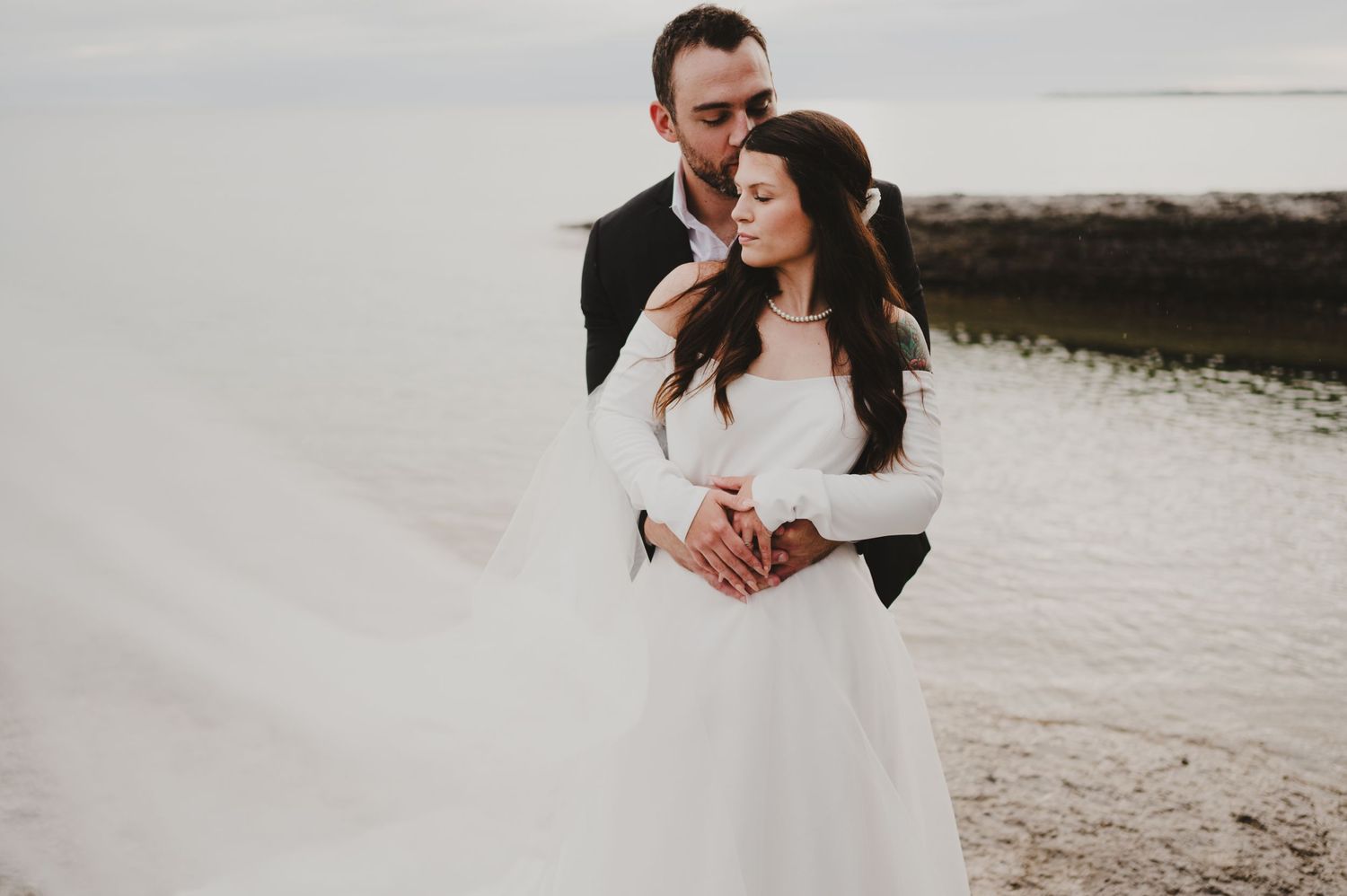 A wedding couple embraces on a rocky beach shore with overcast skies in the background.