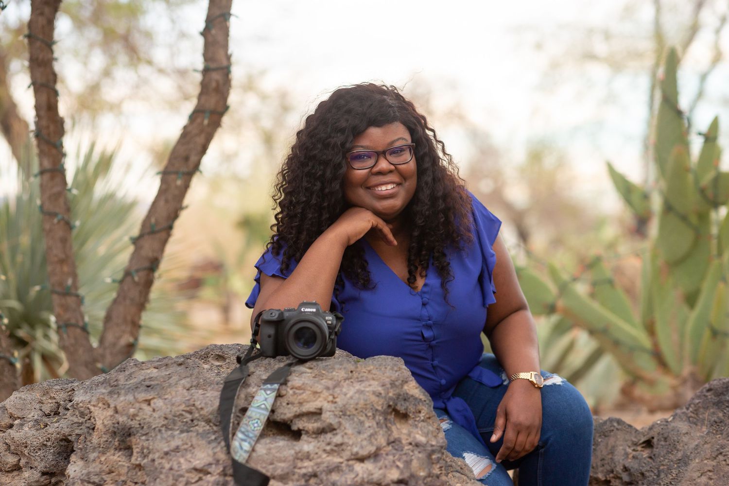 Smiling woman with curly hair sitting on rocks holding a camera in a desert garden setting.