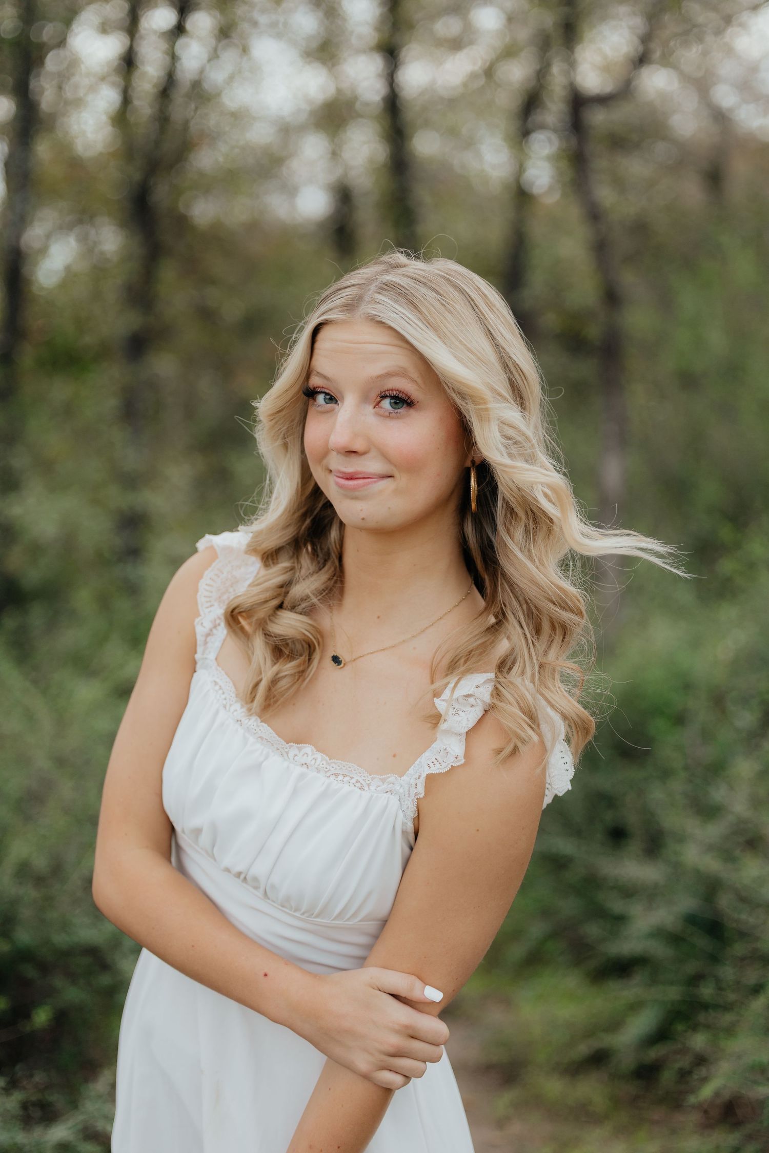 Young blonde woman in white dress posing outdoors among green trees.