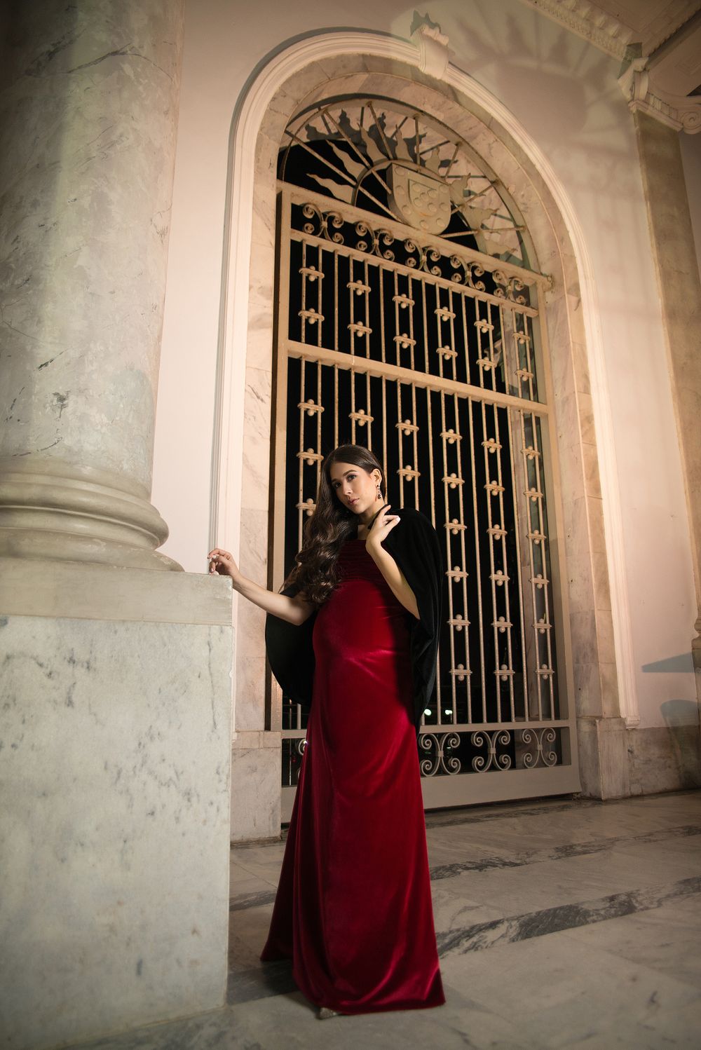Elegant maternity photo in red velvet dress beside ornate iron gate.