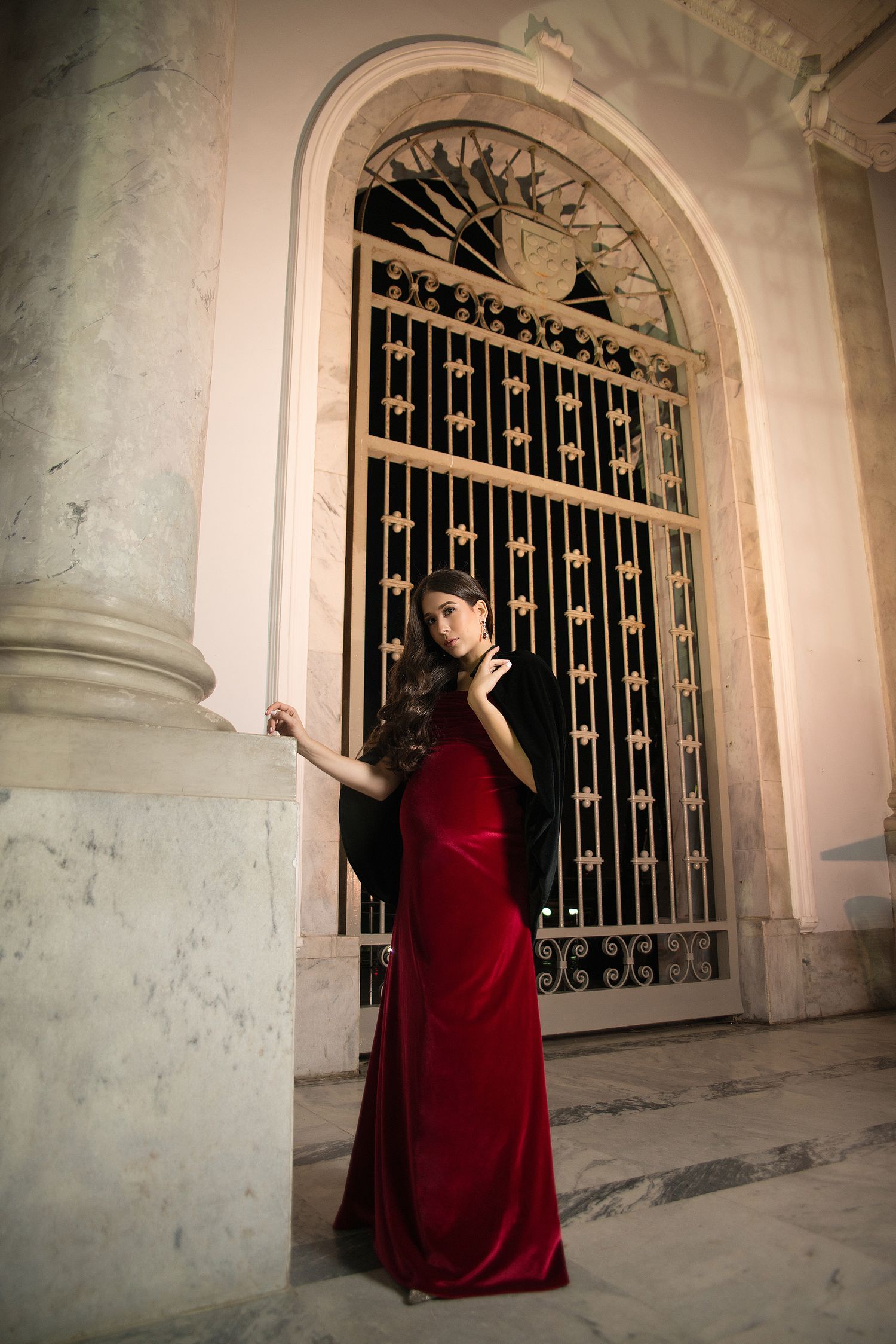 Elegant maternity photo in red velvet dress beside ornate iron gate.