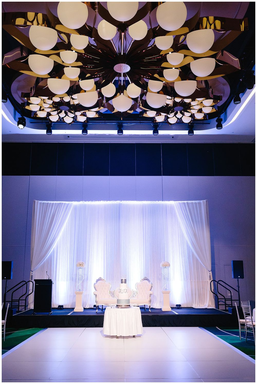 Modern ballroom wedding venue with dramatic ceiling lights and white draped backdrop on stage.