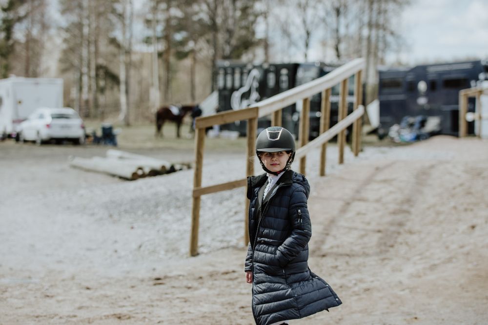 Equestrian in riding gear and helmet standing on dirt path near stables.