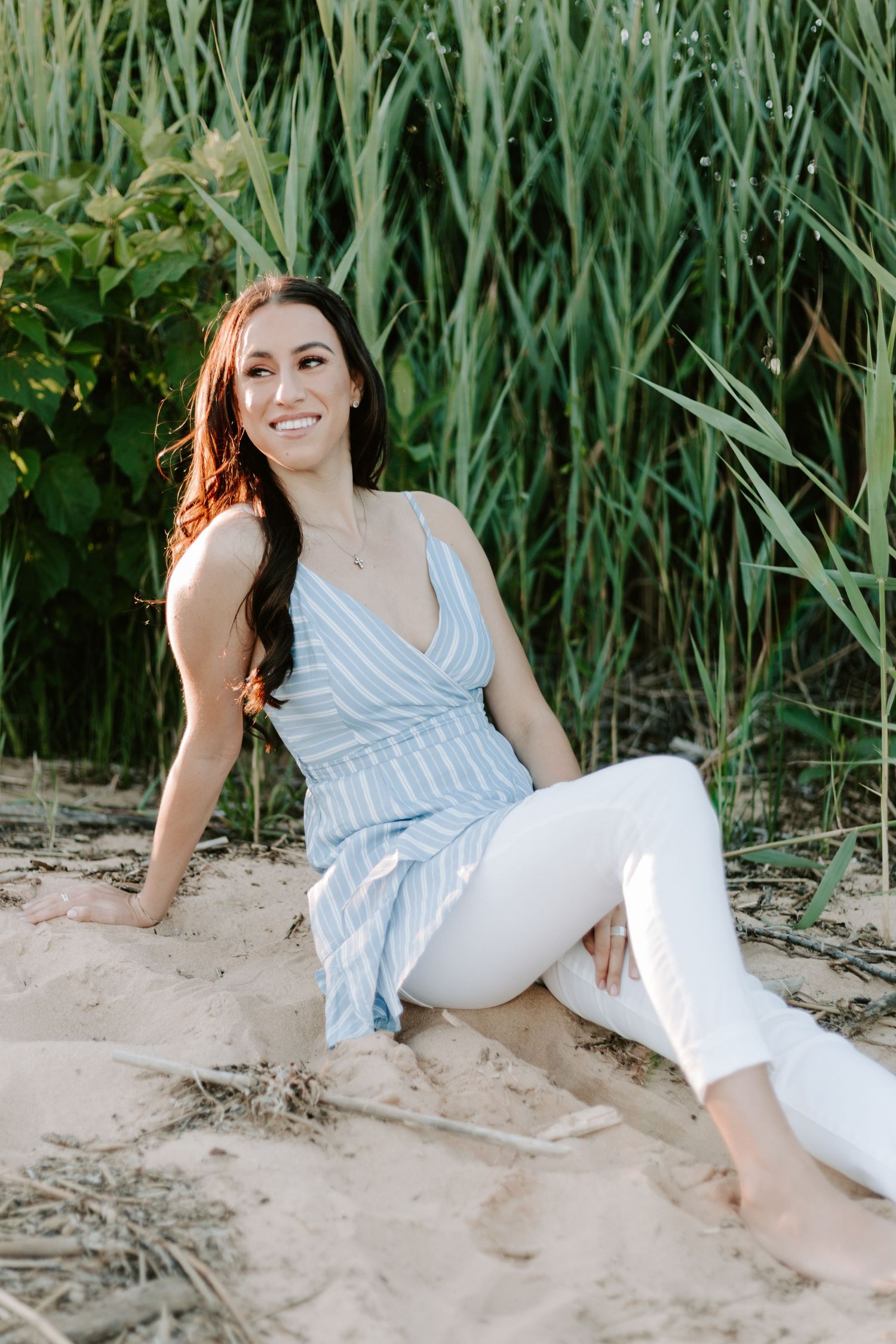 A woman in a blue striped dress sitting on a sandy beach near tall grass on a summer day.