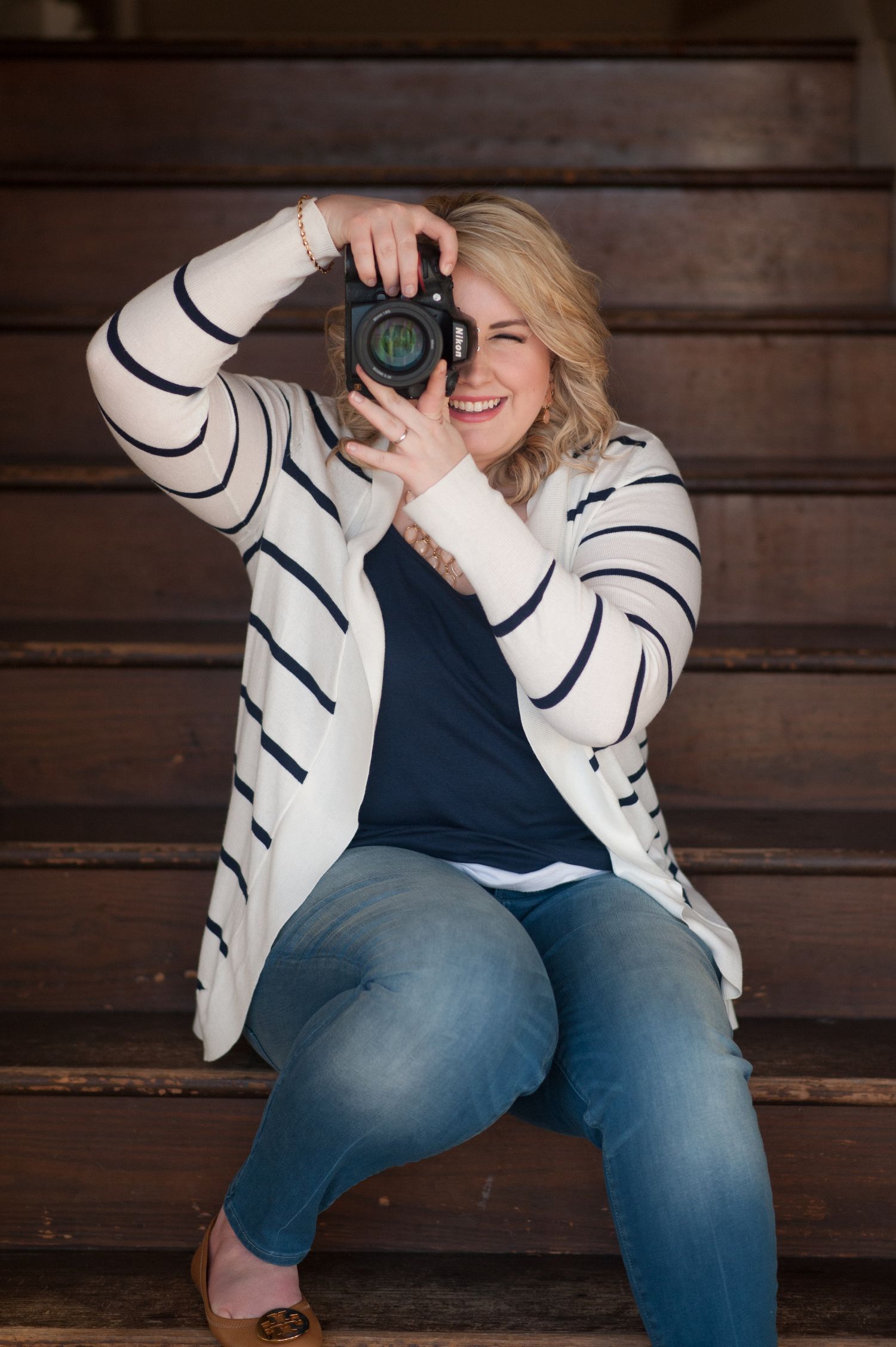 Professional photographer in white striped jacket and jeans sitting on wooden steps with camera.