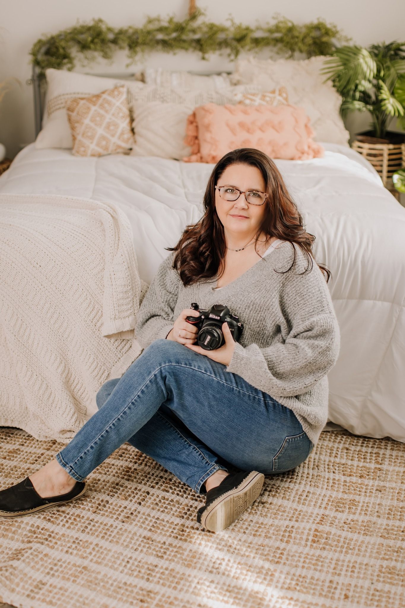 Woman in gray sweater and jeans sits on floor holding camera in front of decorated bed with plants.