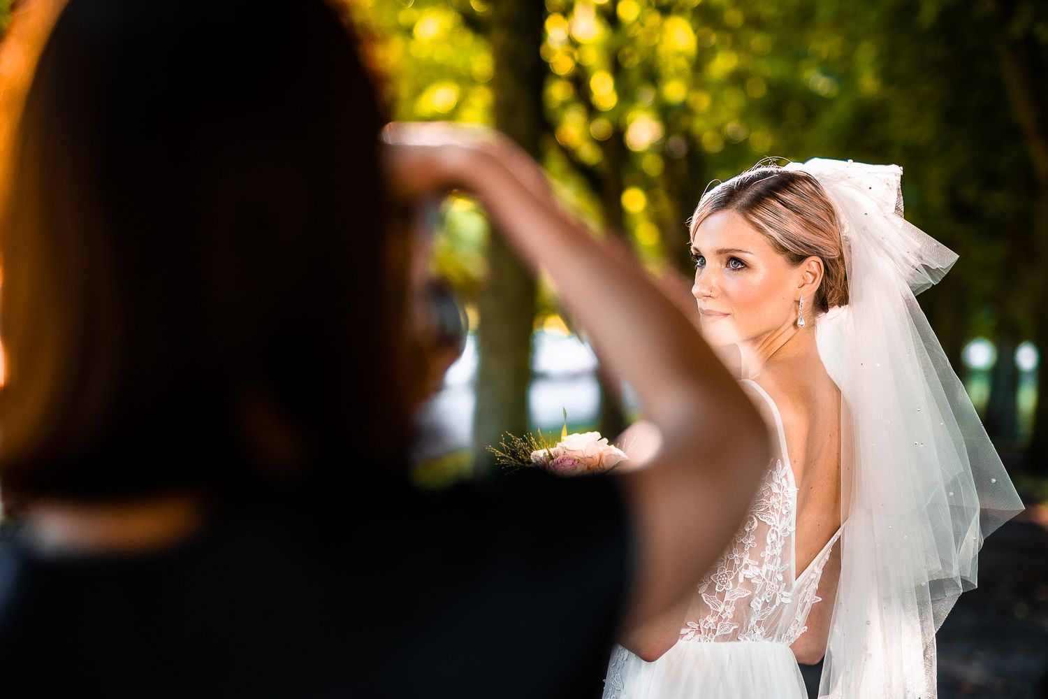 A radiant bride in a lace wedding dress and veil poses outdoors while being photographed in natural lighting.