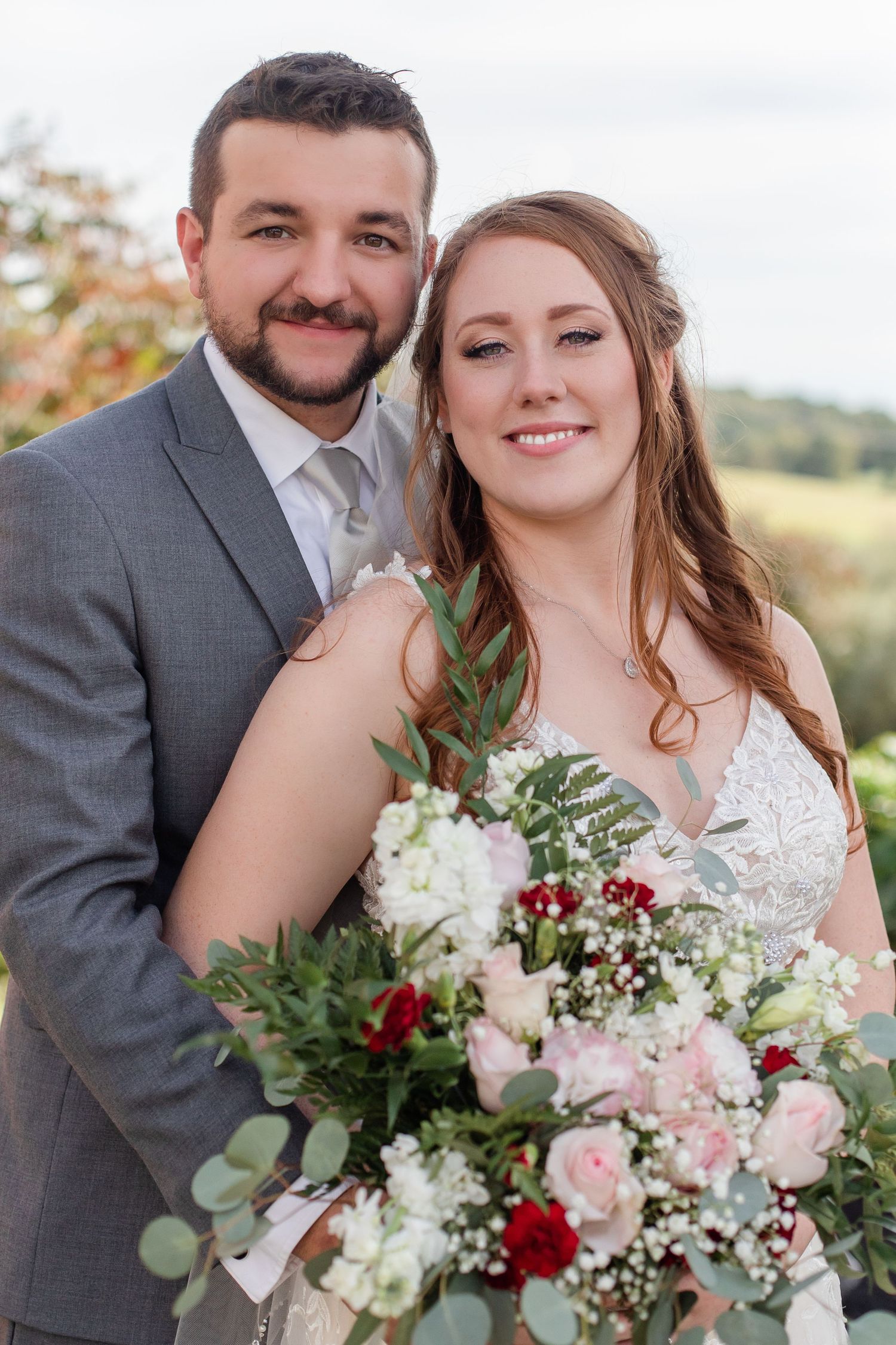 Wedding couple poses with a beautiful bouquet of pink and red flowers with greenery.