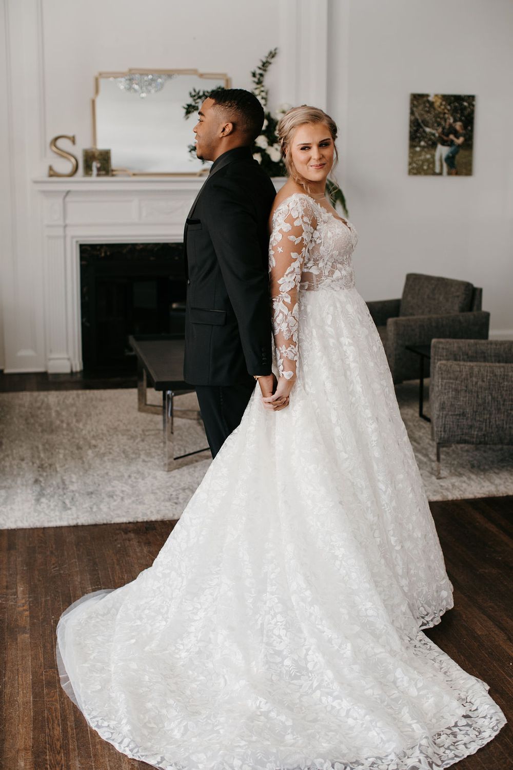 Bride in floral lace long-sleeve ballgown stands back-to-back with groom in black tuxedo indoors.