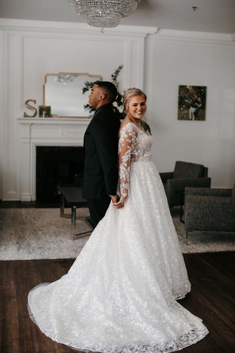 Bride in floral lace ballgown and groom in black tuxedo stand back-to-back holding hands indoors.