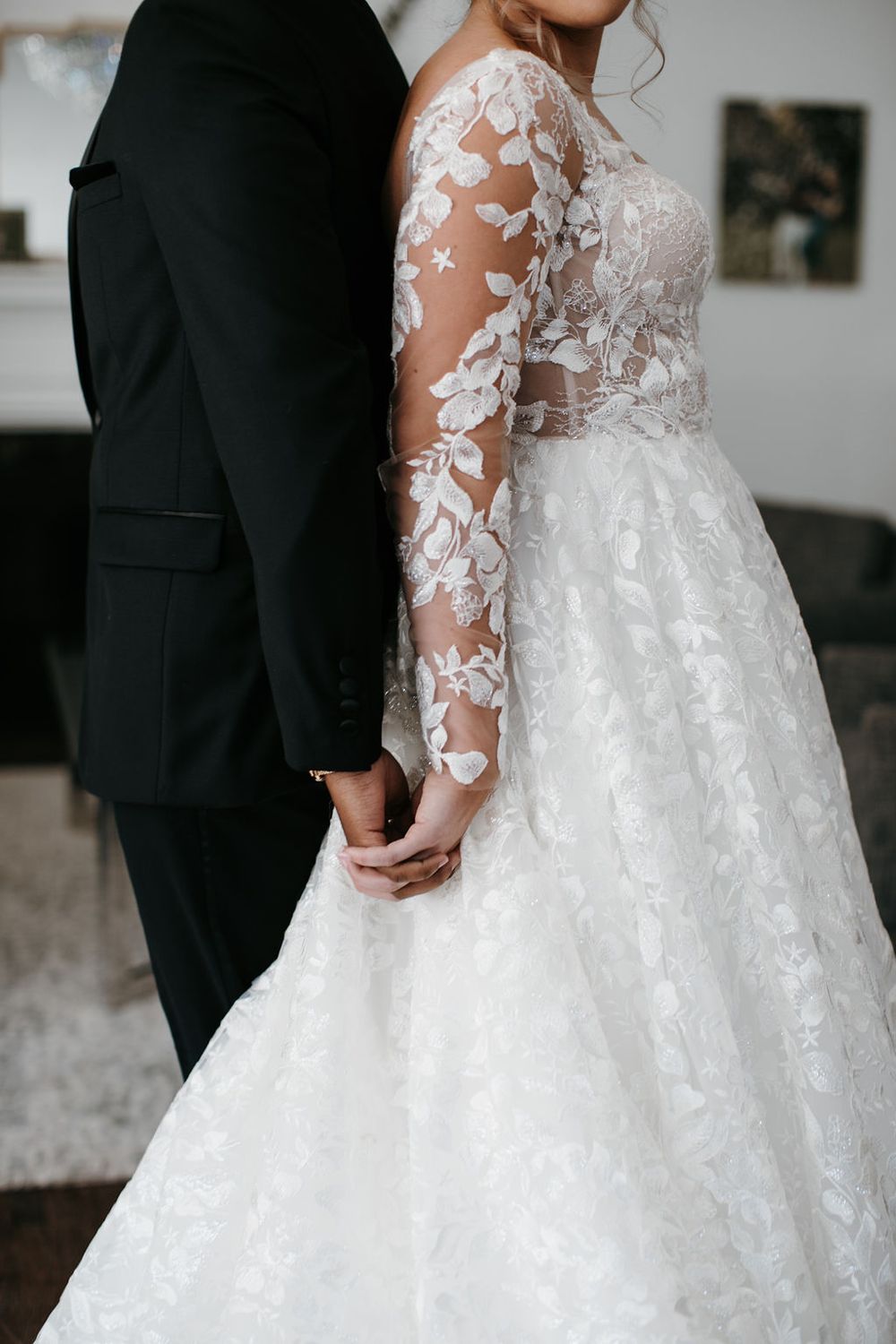Close-up of bride in sheer floral lace long-sleeve wedding gown holding hands with groom in black suit.