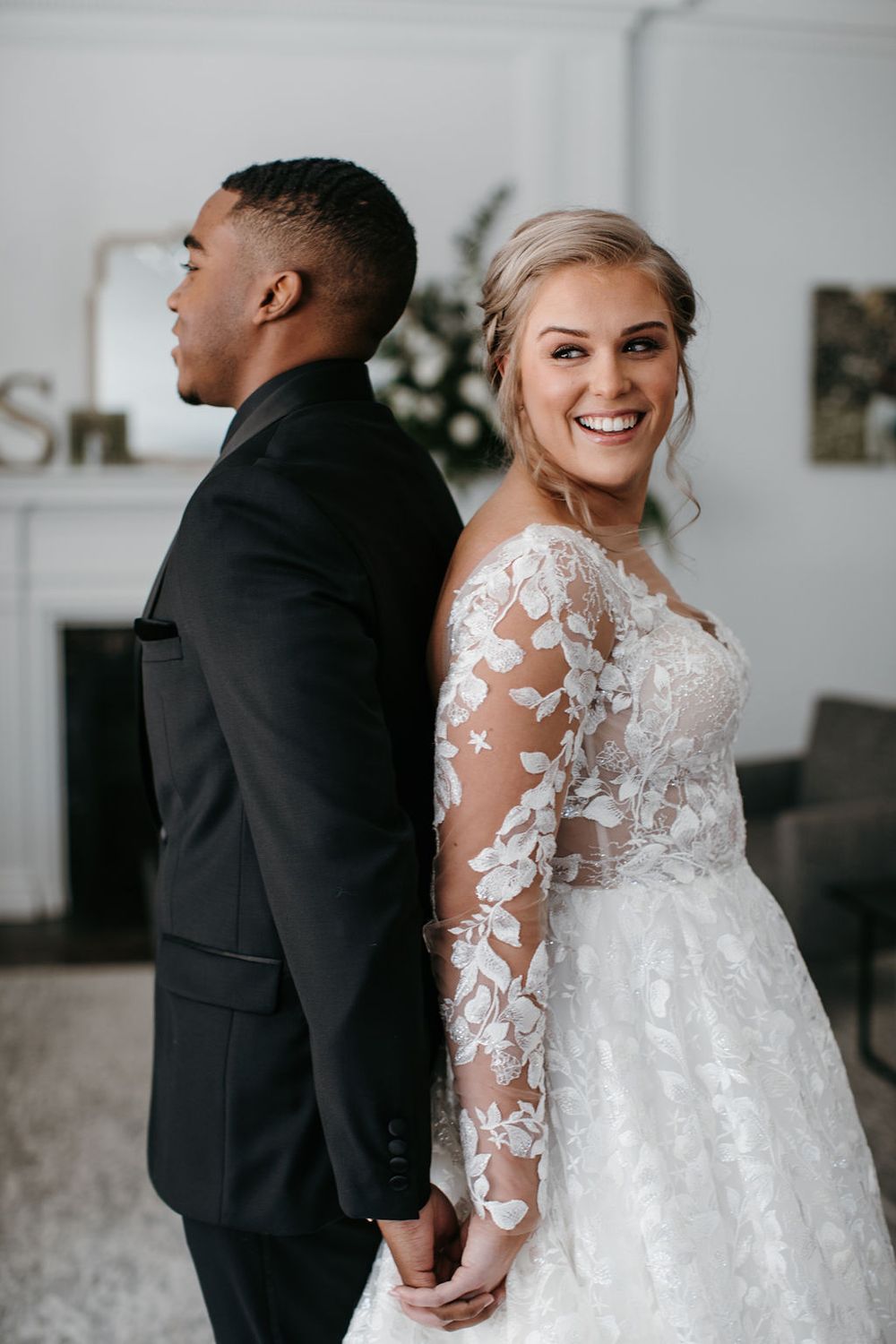 Smiling bride in lace long-sleeve gown poses back-to-back with groom in black tuxedo by fireplace.