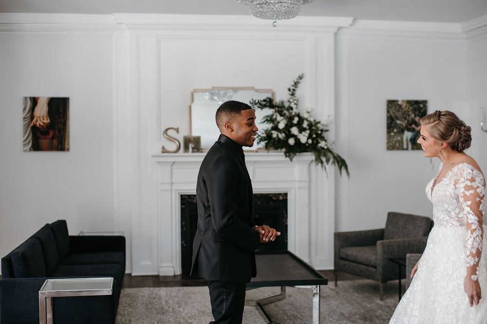 Groom in black tuxedo smiles at bride in lace long-sleeve gown during indoor first look reveal.