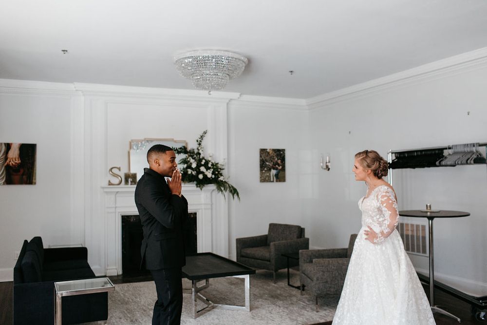 Groom in black tuxedo turns to see bride in lace long-sleeve gown during emotional first look moment.