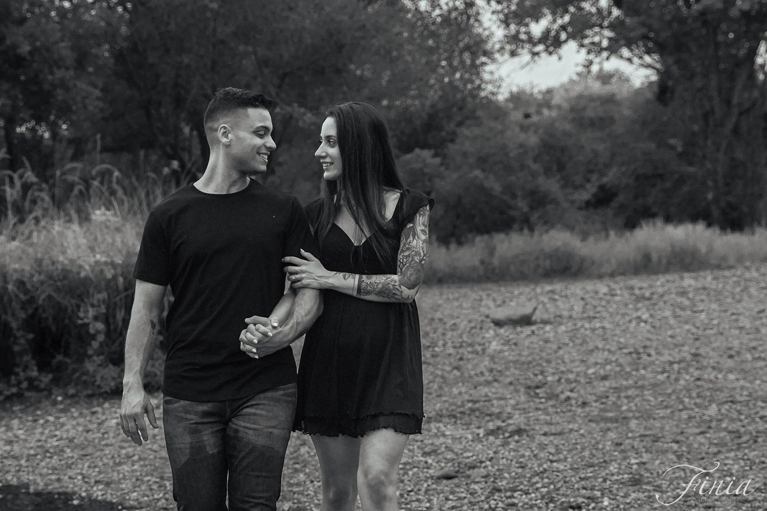 A couple in black clothing shares a tender moment while walking on a dirt path in a black and white photo.