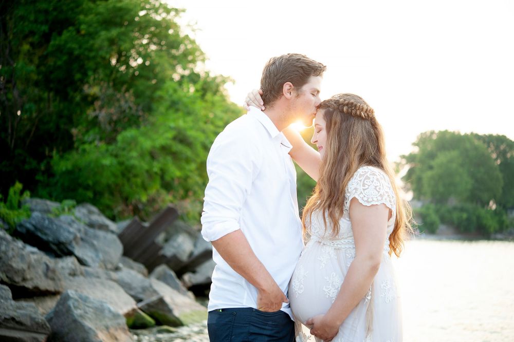 Romantic maternity photo of couple embracing by rocky waterfront at sunset.