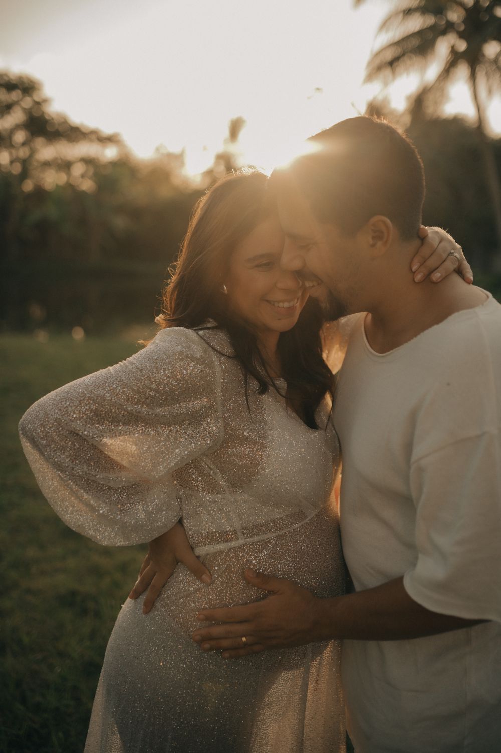 Intimate maternity photo of couple embracing in golden sunset light with palm trees in background.