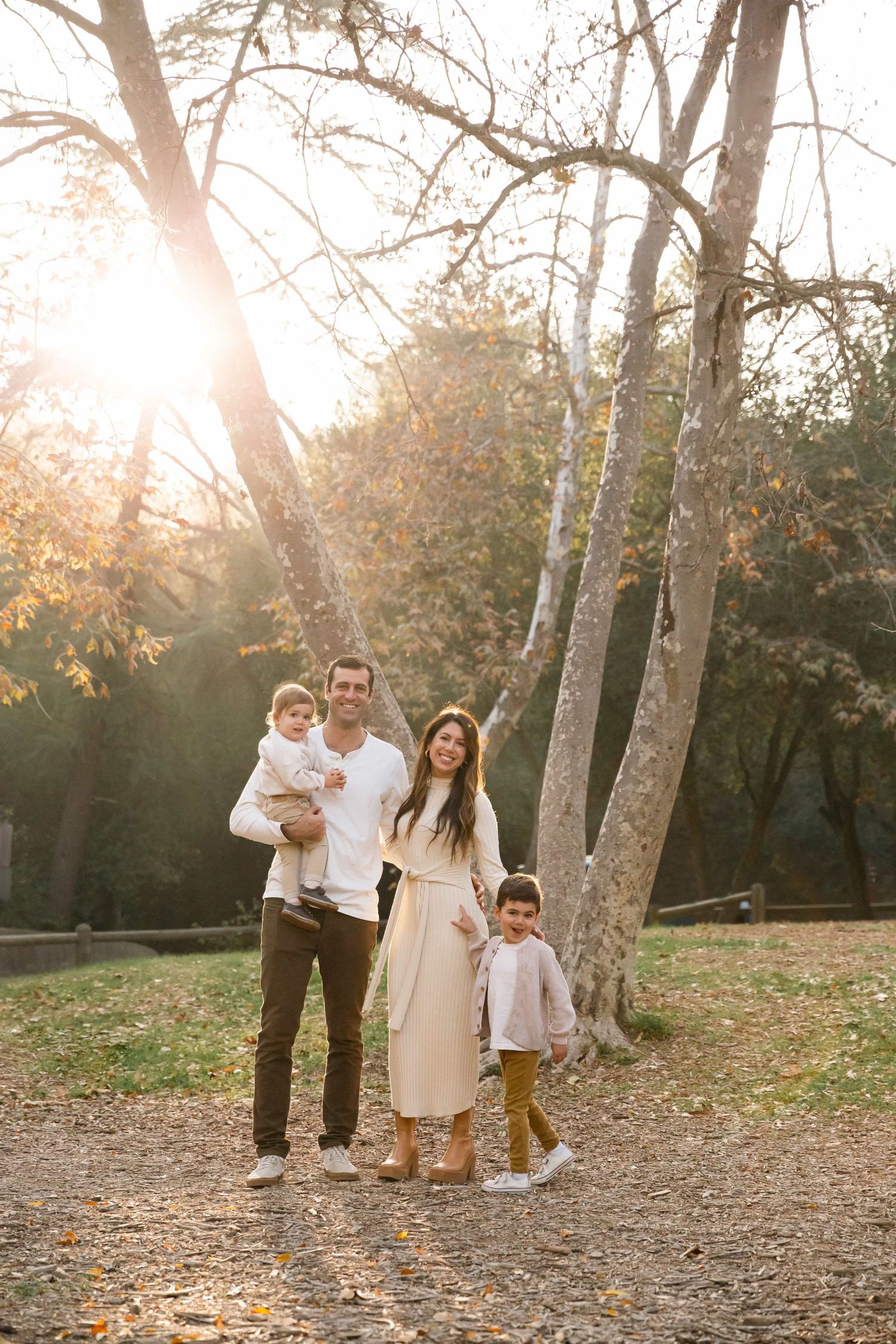 Family of four posing in a sunlit autumn park with tall trees and golden fallen leaves.