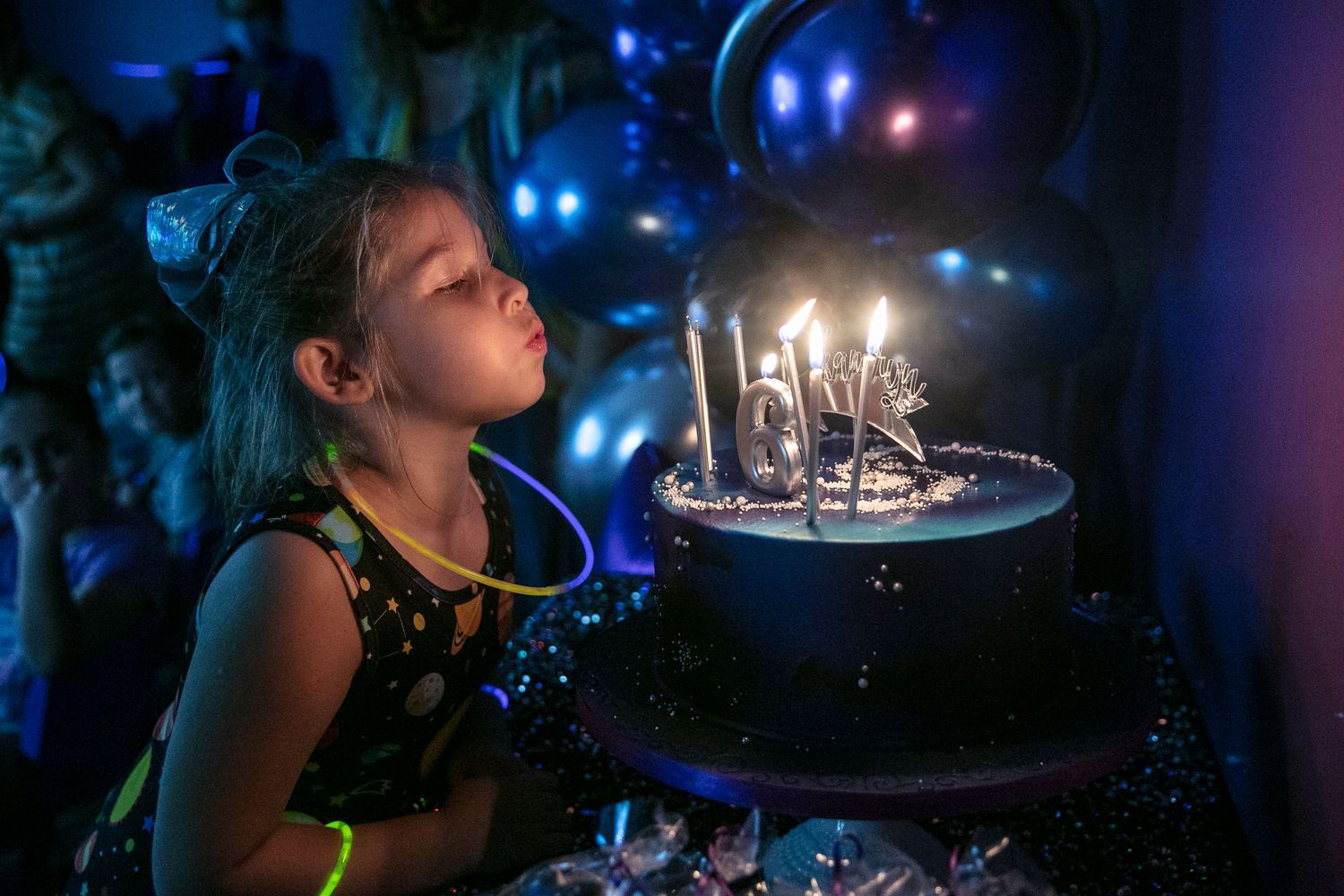 A birthday cake with glowing candles illuminated in a dark room with blue bokeh lights in the background.