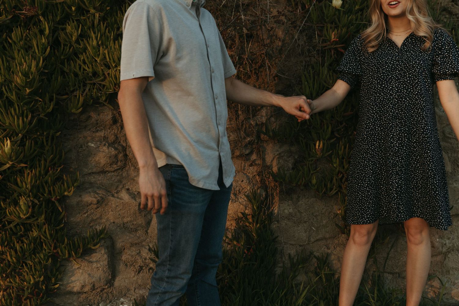 A couple in casual attire standing against a stone wall with greenery.