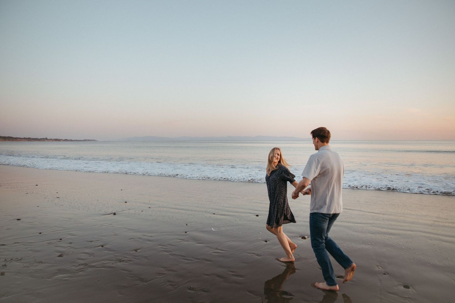 Couple dancing and holding hands on an empty beach at sunset with ocean waves in the background.