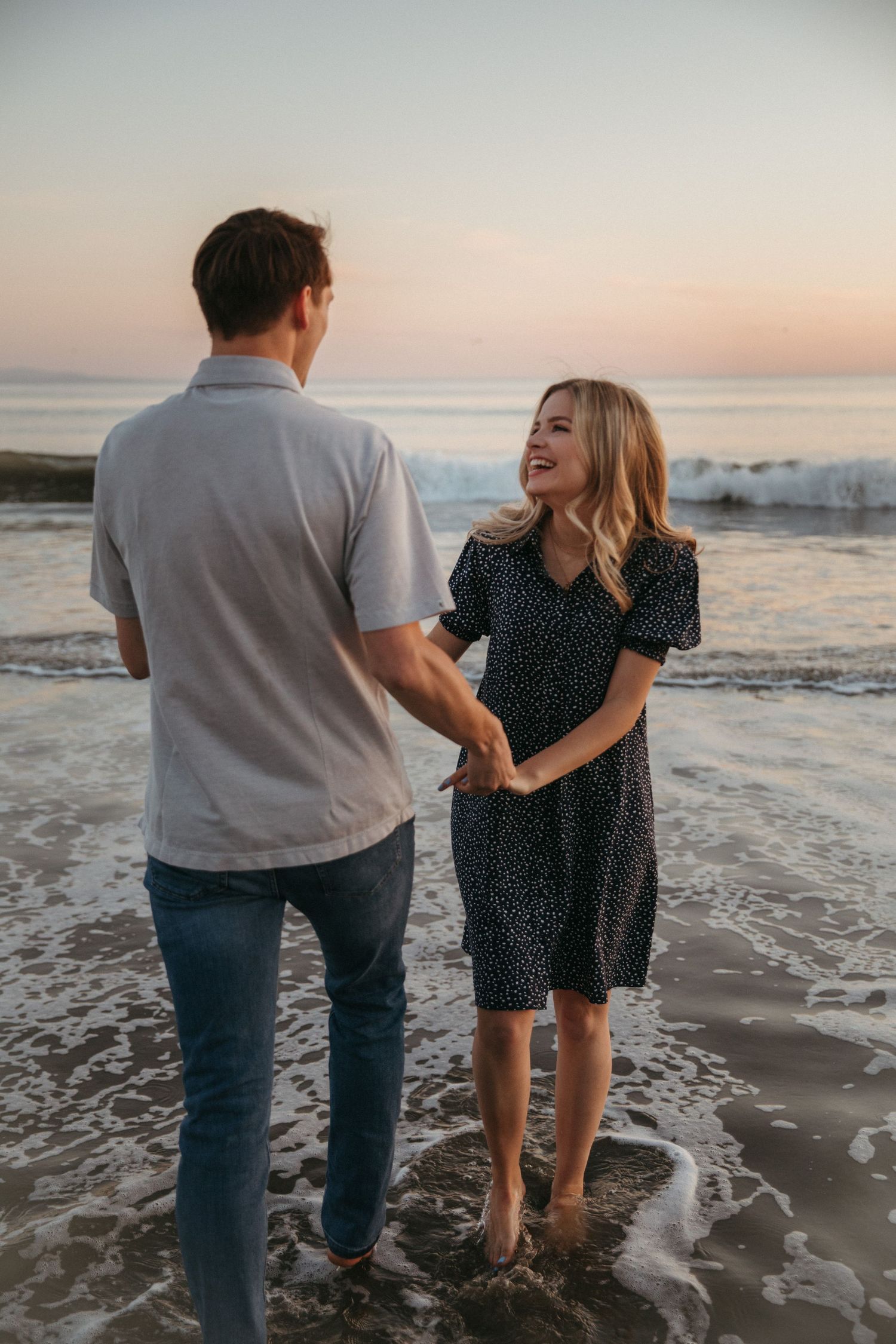 Couple sharing laughter while standing barefoot in shallow ocean waves during golden hour.