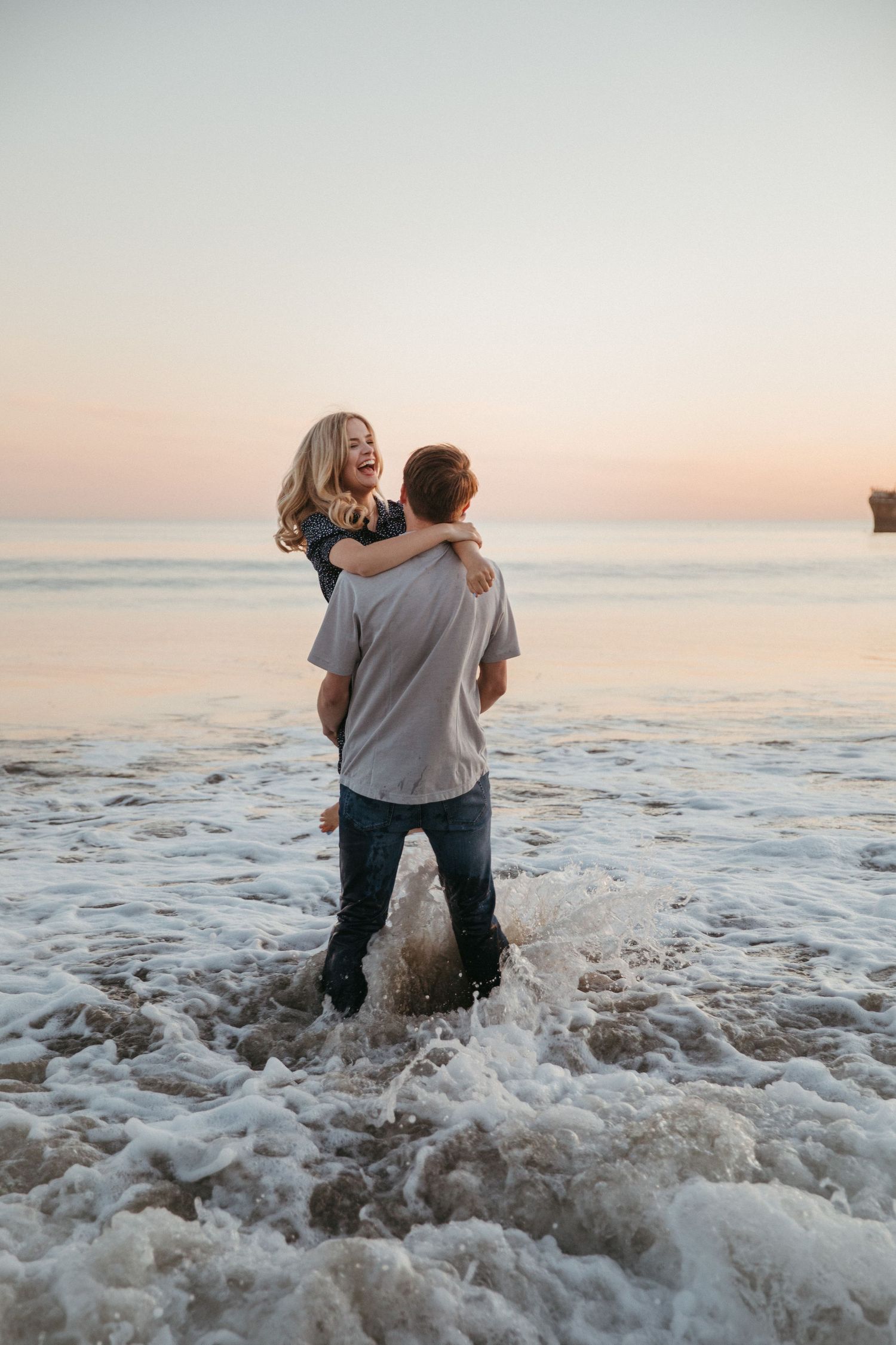Joyful couple playing in ocean waves during sunset with soft pink sky in background.