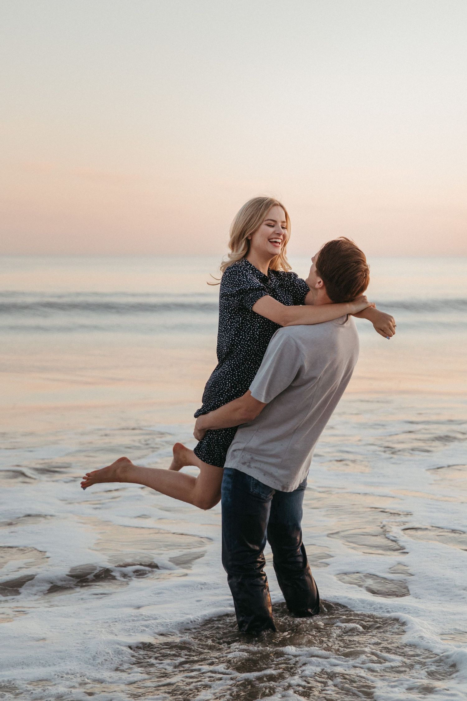 Romantic couple embracing and laughing while standing in gentle ocean waves at sunset.