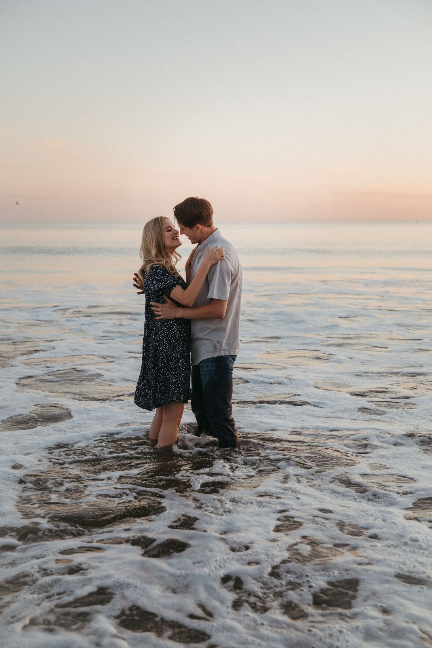Intimate moment between couple standing in ocean waves during sunset with pastel sky.